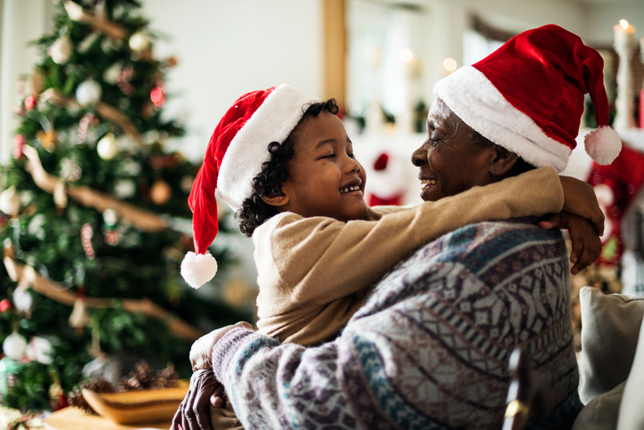 Grandparent and grandchild hugging wearing santa hats in front of a Christmas tree